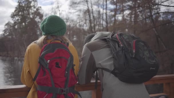 Két turista öltözött kabátok és kalapot állt a hídon, közel a folyó beszélni és élvezni a természetet. Egy fiatal házaspár nyaralását. Barátok töltik a hétvégén a szabadban. — Stock videók