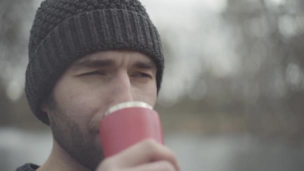 Close up portret van een bebaarde zelfverzekerd glimlachende hipster man in een warme hoed drinken van thee of koffie uit een thermo mok buitenshuis. De man is ontspannen in het weekend in het Park. — Stockvideo