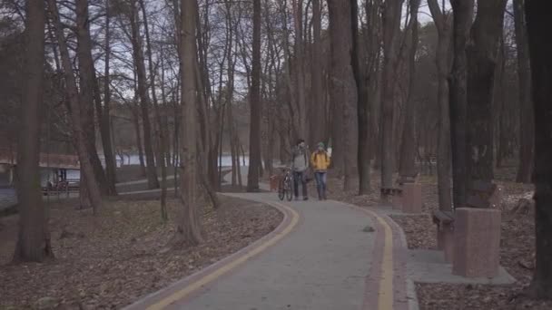 Jovem casal de turistas caminhando ao longo do caminho no parque com mochilas nas costas. O homem leva a bicicleta e a mulher de casaco amarelo brilhante caminha perto. Amantes felizes descansando ao ar livre — Vídeo de Stock