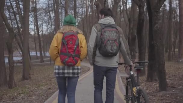 Fiatal turisták séta a parkban, hátizsákok a hátukon. Az ember vezeti a kerékpárt, és nő a fényes sárga kabát sétál közelében. Boldog szerelmesek pihenés kint. — Stock videók