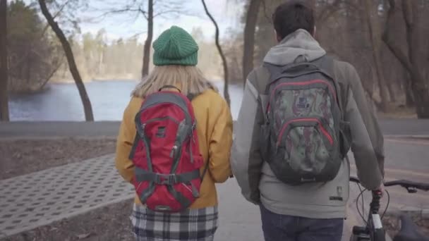 Rear view on young tourists walking in the park with backpacks on their backs. The man leads the bicycle and woman in bright yellow jacket walks near. Happy lovers resting outdoors. — Stock Video
