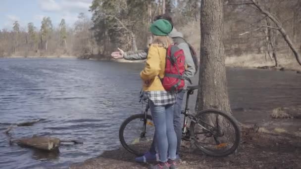 Fiatal pár turista állt a folyóparton a hátizsákok a hátukon. A férfi tartja a kerékpáros és a nő a világos sárga kabát álló közelében. Szerelmesek többi szabadban. Oldalnézet — Stock videók