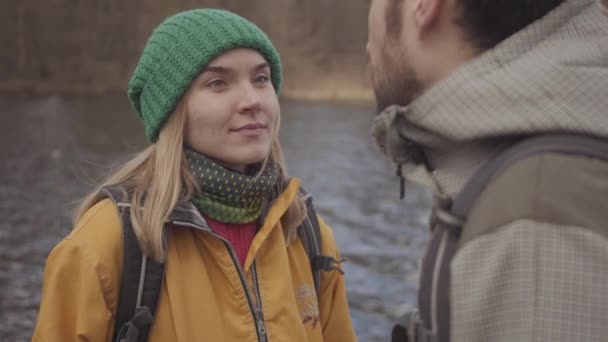 Portrait of happy young couple of tourists standing on the river bank with backpacks on their backs. Bearded man and pretty woman in yellow jacket talking and laughing. Lovers rest outdoors — Stock Video