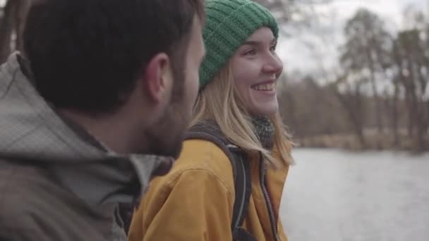 Közelről portré fiatal pár turista hátizsákok ül a padon, a tavaszi parkban, a folyóparton. A szakállas férfi és a sárga kabát nő beszél. Szerelmesek pihenés kint — Stock videók