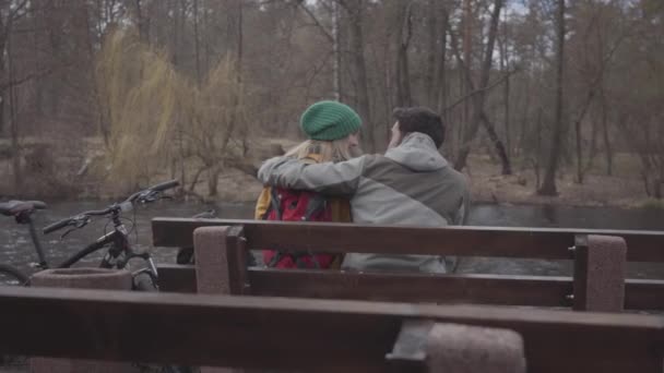 Ein junges Touristenpaar mit Rucksack sitzt auf der Bank im Frühlingspark am Flussufer. der bärtige Mann und die bärtige Frau in der gelben Jacke im Gespräch. Liebhaber ruhen sich im Freien aus — Stockvideo