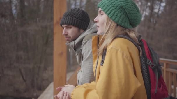 Retrato de la joven y confiada pareja de turistas parados en el puente con mochilas en la espalda. Hombre barbudo y mujer bonita con chaqueta amarilla hablando y sonriendo. Los amantes descansan al aire libre — Vídeo de stock