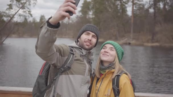 Porträtt av unga par resenärer tar selfie medan du står på en bro mot bakgrund av en liten flod. Unga människor är klädda i jackor och hattar. Semester framgångs rik oberoende — Stockvideo