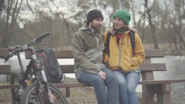 Um jovem casal de turistas com mochilas sentadas no banco do parque primaveril na margem do rio. O homem e a mulher barbudos de casaco amarelo a falar. A bicicleta que está perto. Os amantes descansam ao ar livre — Vídeo de Stock