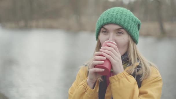 Portret van mooie jonge vrouw in groene hoed en gele jas drinken van thee of koffie uit de Thermos in het Park op zoek in de camera close-up. Positieve dame rust buitenshuis. — Stockvideo