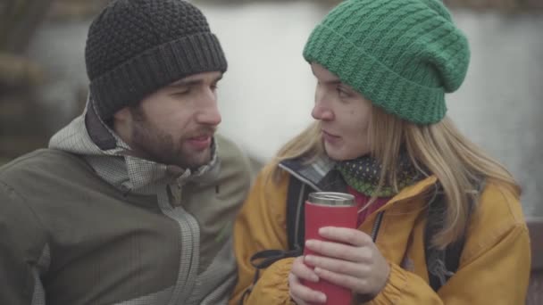 Портрет молодой пары в теплых пальто, разговаривающей на улице. Женщина в жёлтой куртке и зелёной шляпе пьёт чай или кофе из термоса. Любовники улыбаются — стоковое видео