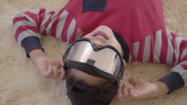 Küçük afro-amerikan çocuk bej kabarık halı üzerinde gözleri üzerinde paintball gözlük ile yerde yatan portre kadar kapatın. Komik çocuk eğlenir. Diğer mutlu çocuklar.