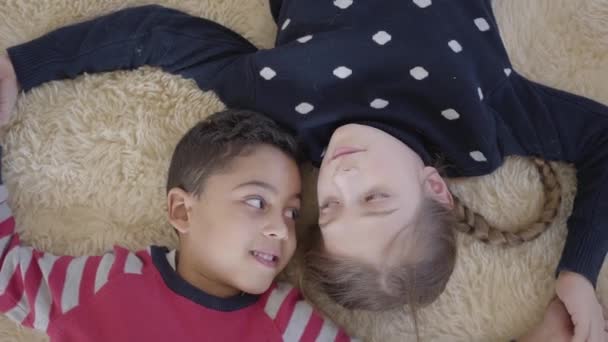 Retrato bonito menino afro-americano e menina caucasiana loira deitada no chão no tapete fofo bege e olhando um para o outro com amor. Vista superior. Resto de crianças felizes . — Vídeo de Stock