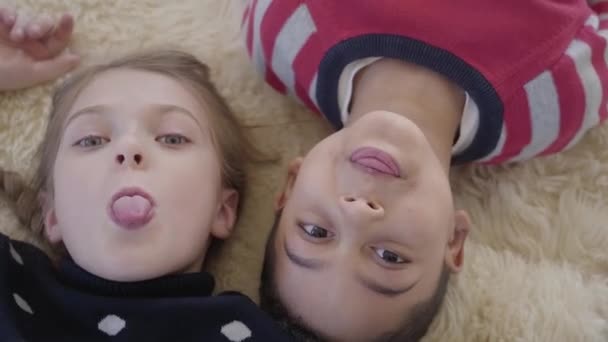Close up portrait cute african american boy and blond caucasian girl lying on the floor on the beige fluffy carpet and looking in camera showing their tongues. Leisure of funny kids. Top view. Rest of — Stock Video