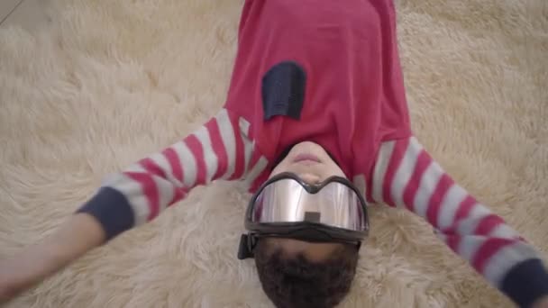 Närbild porträtt av bedårande afrikansk amerikansk pojke som ligger på golvet på den beige fluffiga mattan med skidglasögon på hans ögon närbild. Rolig pojke har ett roligt. Övriga glada barn. — Stockvideo