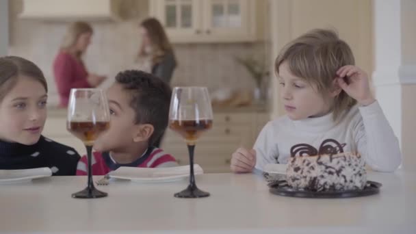 Portrait three funny kids sitting at the table with small cake and wine  glasses with juice. Two caucasian girls and african american boy have fun  together — Stock Video © photo_oles #257720062