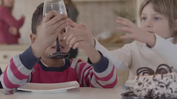 Dois miúdos sentados à mesa em casa. Menina loira caucasiana e menino afro-americano se divertir juntos. O rapaz que dá o copo com o suco à amiga. Lazer de dois miúdos bonitos engraçados de raças diferentes — Vídeo de Stock