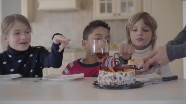 Tres niños en la mesa con tarta pequeña y vasos de jugo. La mano de una mujer corta el pastel y pone un pedazo al plato de los niños. Dos chicas caucásicas y un chico afroamericano esperando sus dulces — Vídeos de Stock