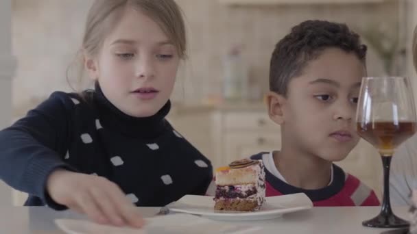 Three kids at the table with small cake and juice glasses. Hand of a woman cuts the cake and puts a piece to boys plate. Two caucasian girls and african american boy waiting for their sweets — Stock Video