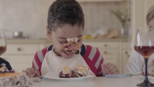 Three kids have a fun together sitting at the table. There is the cake smeared on boy face. Two caucasian girls and african american boy laughing. Leisure of cute kids of different races — Stock Video