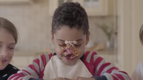 Los niños se divierten juntos sentados en la mesa. Ahí está la torta untada en la cara del niño, el niño amasando la torta en el plato. Dos chicas caucásicas y un chico afroamericano sonriendo. Ocio de niños de diferentes razas — Vídeos de Stock