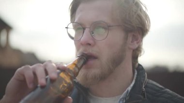 Yakından portre yakışıklı sakallı adam gözlük bira içme ve açık havada içecek zevk ile. Adam kameraya bakarak şişeden bira tadı. Yavaş çekim.