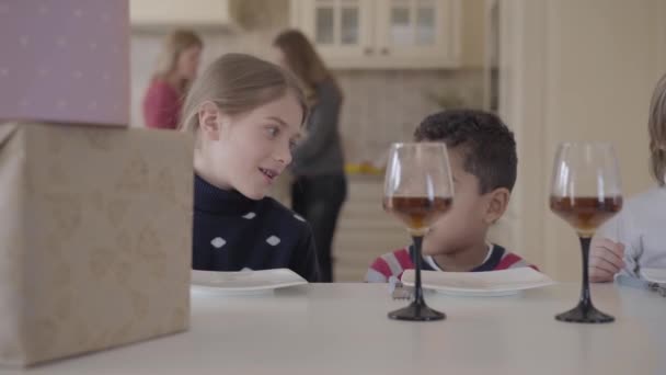 Retrato três crianças engraçadas sentadas à mesa com bolo pequeno e copos de vinho com suco. Duas garotas caucasianas e o garoto afro-americano se divertem juntos — Vídeo de Stock