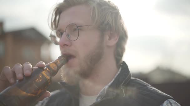 Портрет блондина, пьющего пиво на улице, отводящего взгляд. Бородатый мужчина в очках наслаждается алкогольным напитком, волосы трепещут на ветру. Медленное движение — стоковое видео