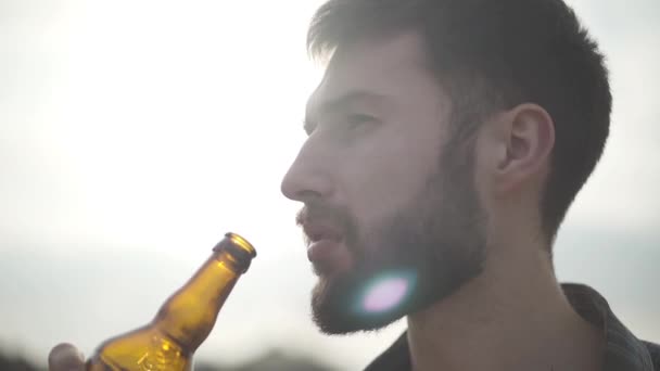 Портрет привлекательный бородатый мужчина, пьющий пиво и наслаждающийся напитками на открытом воздухе. Парень пробует пиво из бутылки, глядя в камеру. Медленное движение . — стоковое видео
