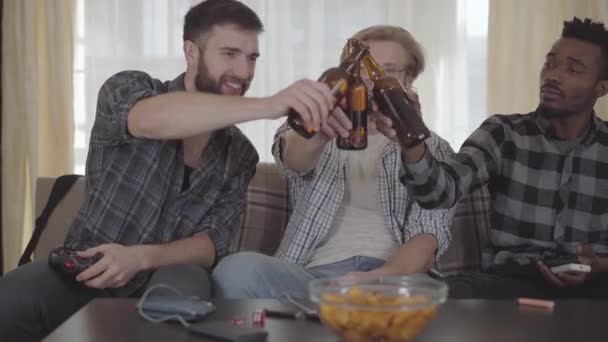 Portrait deux hommes caucasiens et un homme afro-américain assis sur le canapé. Trois hommes assis ensemble à la maison buvant de la bière. Les amis cliquent leurs bouteilles. Un homme s'étouffa, tout le monde riait — Video