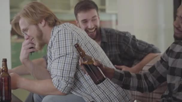 Dos caucásicos y un afroamericano sentados en el sofá. Tres hombres sentados juntos en casa bebiendo cerveza. Los amigos rompen sus botellas. Un hombre se ahogó, todos riendo — Vídeo de stock