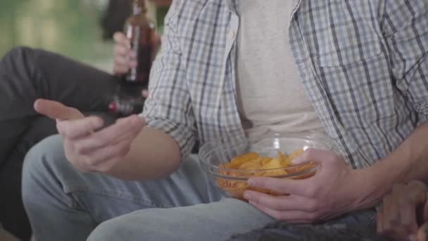 Друзья отдыхают, пьют пиво и едят чипсы сидя на диване в закрытом помещении . — стоковое видео