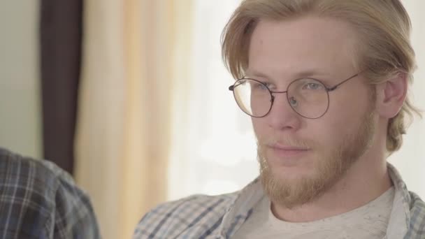 Portrait of the confident blond man with blue eyes drinking beer indoors looking away. Bearded man in glasses enjoying his alcohol drink sitting at home on the sofa — Stock Video