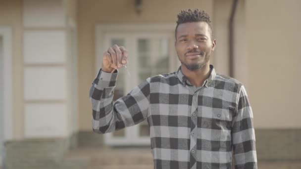 Handsome african american man in checkered shirt standing in front of a big house holding keys in his hand and smiling. The man just bought a house. The guy stands near the new home — Stock Video