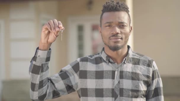 Portrait confiant bel homme afro-américain en chemise à carreaux debout devant une grande maison tenant les clés dans sa main et souriant. Il vient d'acheter une maison. Le gars se tient près de la nouvelle maison — Video