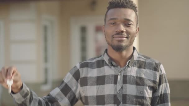 Portret van knappe Afro-Amerikaanse man in geruit overhemd dat de sleutels voor een groot huis demonstreert. De man draait en loopt naar de deur van zijn nieuwe huis. De man net gekocht huis — Stockvideo