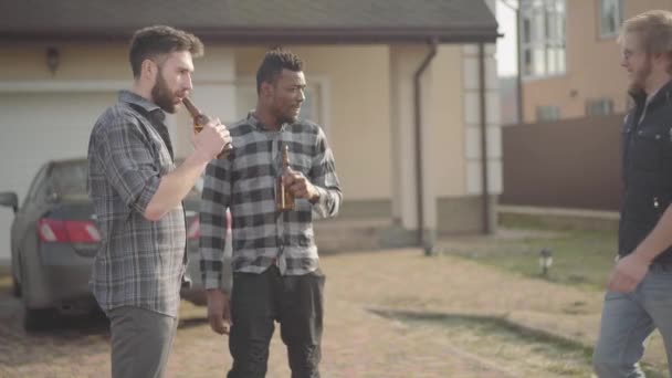 大きな家の前に立つ2人の白人とアフリカ系アメリカ人の男性がビールを飲んでいます。三番目の友人が来て、男性はお互いに挨拶し、抱き合う。古い友人は一緒に楽しみを持っています — ストック動画