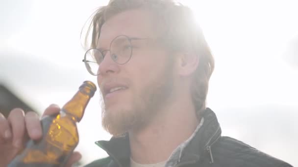 Retrato do loiro a beber cerveja ao ar livre a desviar o olhar. Homem barbudo de óculos desfrutando de sua bebida alcoólica, cabelo balançando ao vento . — Vídeo de Stock