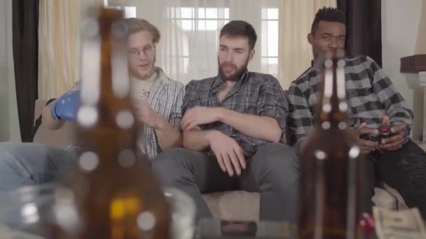 Friends watching television sitting on the couch. On the table are empty beer bottles. The guys are fooling around. Bearded young man wearing a blue boxing glove and jokingly boxing with friends. — Stock Video