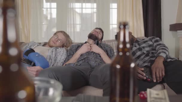 Potret dua pria Kaukasia dan satu pria afrika yang tidur di sofa setelah menonton tinju di TV. Botol bir kosong dan mangkuk keripik berada di atas meja . — Stok Video