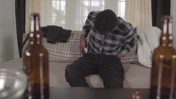 Όμορφος Αφροαμερικανός άνθρωπος που ελέγχει την τσέπη του καθισμένος στον καναπέ και βρίσκει μόνο ένα δολάριο. Τα θολά άδεια μπουκάλια μπύρας είναι στο τραπέζι. Πρωί μετά το μεθύσι. Ανθυγιεινή ζωή — Αρχείο Βίντεο