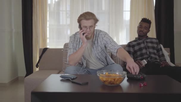 Bonito homem barbudo sentado no sofá falando por celular, enquanto seu amigo afro-americano está perto de desviar o olhar. Os dados e a tigela de batatas fritas estão na mesa. Rapazes vão divertir-se em casa — Vídeo de Stock