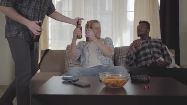 Dos hombres caucásicos y afroamericanos sentados en el sofá. El tercer amigo viene con botellas de cerveza y se las da a los chicos. Los hombres se saludan. Viejos amigos van a ver fútbol en casa — Vídeos de Stock