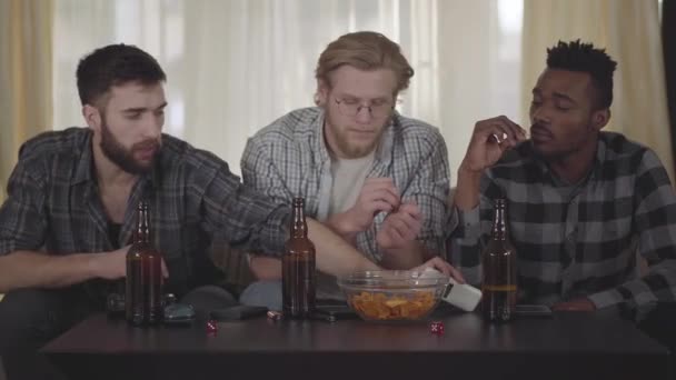 Dos caucásicos y un afroamericano descansando juntos en casa, divirtiéndose. Botellas de cerveza y patatas fritas en la mesa. El tipo cambia los canales de TV usando el control remoto. Viejos amigos descansan juntos — Vídeos de Stock