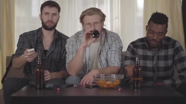 Portret trzech szczęśliwych przyjaciół siedzi w domu na cauch, patrząc TV, picie piwa z frytkami, o rozmowę wideo do kogoś. — Wideo stockowe
