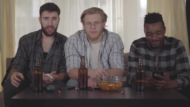 drei befreundete glückliche Männer, die im Haus auf der Couch sitzen, fernsehen, Bier mit Chips trinken, einen Videoanruf mit jemandem haben.