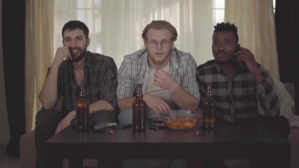 Portrait de trois jeunes hommes amis assis à la maison sur la marche, regarder la télévision, boire de la bière avec des jetons, regarder le match de football et très unset et déçu par le jeu — Video