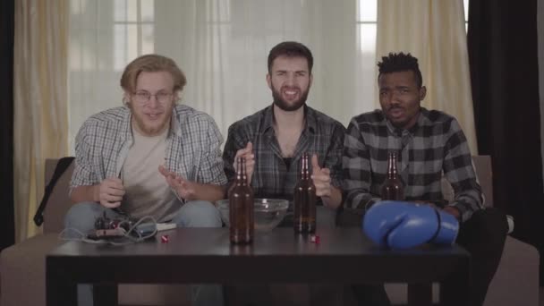 Dva šťastní bělošští a jeden afričtí američtí muži odpočívají doma a dívají se na box v televizi. pivní láhve, prázdná miska na hranolky a boxerská rukavice na stole — Stock video