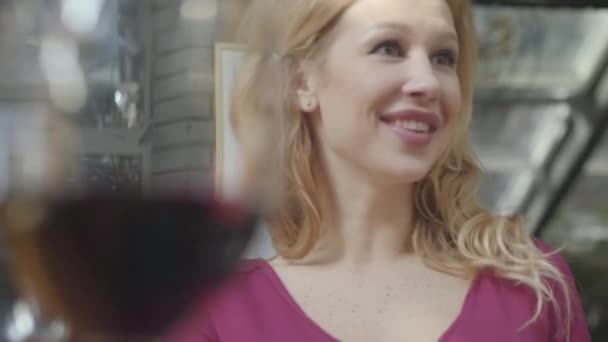 Portret van mooie elegante blonde vrouw met krullend haar praten met vriend op de achtergrond van wazig glas rode wijn. De dame is ontspannen, ze is blij en glimlachend — Stockvideo