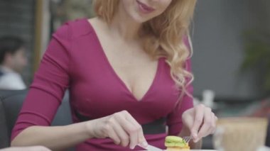 Kıvırcık saçlı çekici sarışın kadın restoranda bir macaroon ile pasta yeme yakın. Derin dekolte ile sıcak bayan onu tatlı sahiptir. Kamera yukarı hareket ediyor