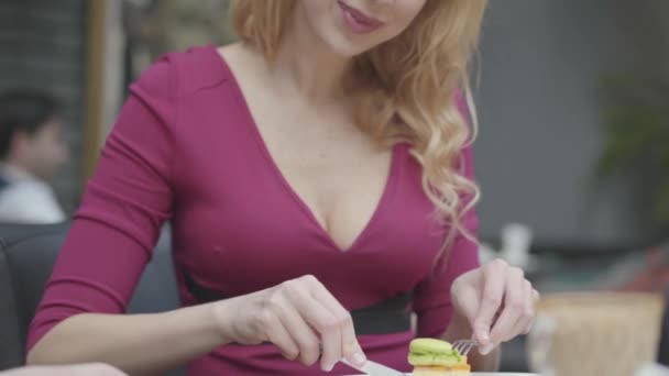 Belle femme blonde avec des cheveux bouclés manger gâteau avec un macaron dans le restaurant de près. Dame chaude avec un décolleté profond aime son dessert. La caméra remonte — Video